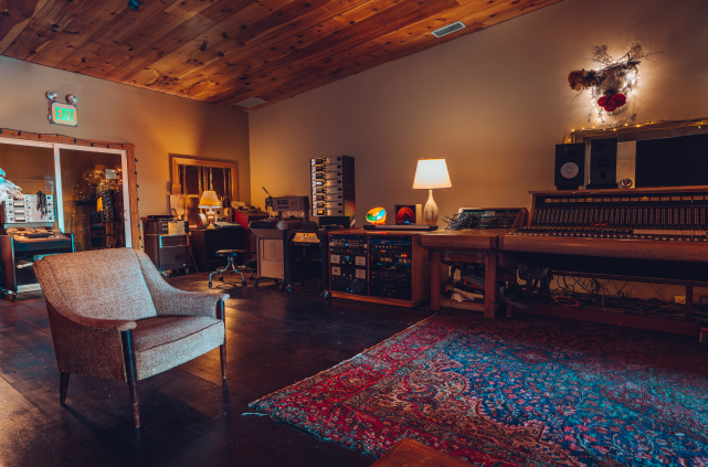 Large room with sound and recording equipment along the wall, a cushioned chain and colorful rug near the middle.
